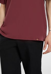 Muž v burgundském tričku s krátkými rukávy a černých kalhotách, s malou logovkou na lemu trička a viditelným tetováním na pravém zápěstí.