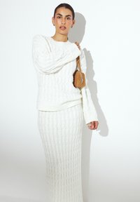 Maglione bianco lavorato a maglia e gonna lunga coordinata con un motivo a treccia. La modella tiene una piccola borsa a tracolla color beige con cerniera.