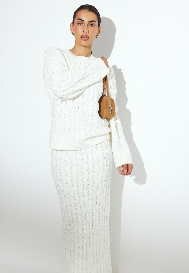 Maglione bianco lavorato a maglia e gonna lunga coordinata con un motivo a treccia. La modella tiene una piccola borsa a tracolla color beige con cerniera.