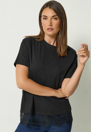 Donna con capelli castani lisci che indossa una maglia nera a maniche corte con dettagli in pizzo e jeans blu, in piedi davanti a uno sfondo chiaro.