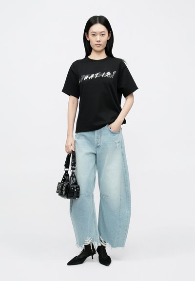 T-shirt de algodão preta com estampa gráfica, combinada com jeans largos azul claro desgastados. Acessórios incluem uma pequena carteira preta.