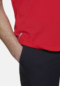 Rode, gestructureerde sportshirt met een zijzoom voorzien van een blauwe en witte accent, gecombineerd met donkere broek. De hand is in de zak geplaatst.