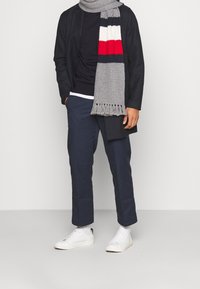 Schwarze Jacke über einem dunklen Hemd, marineblaue Hose und weiße Sneakers; ein grauer Schal mit rot-weiß-blauen Streifen, verziert mit Fransen.