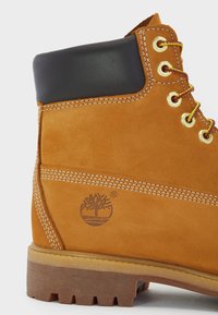 Bruine suede laars met een zwarte leren kraag, gouden oogjes en een rubbersole. Bevat gestikte details en een boomlogo aan de zijkant.