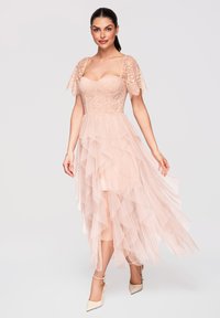 Femme portant une robe à corsage en dentelle rose clair avec des manches flottantes et une jupe en tulle superposée, associée à des talons crème à bride cheville.