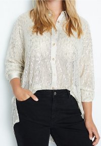 Doorzichtige crèmekleurige blouse met pailletten, knoopsluiting aan de voorkant, lange mouwen en zijslits, gecombineerd met een zwarte broek. Glanzende textuur met subtiel patroon.