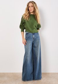 Pullover verde a coste con maniche a sbuffo abbinato a jeans in denim blu a vita alta e gamba larga. Dettagli notevoli includono cuciture arancioni.