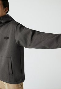 Sweatshirt gris avec un col montant, manches longues et une poche kangourou. Comprend un tissu texturé et un logo brodé noir.