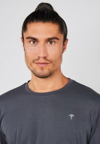 T-shirt gris à manches longues avec un col rond et un petit logo blanc sur le côté gauche de la poitrine. Texture lisse et design ajusté.
