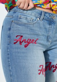 Jeans de mezclilla azul claro con bordado de terciopelo rojo "Ángel" en el muslo, diseño de cinco bolsillos y costuras contrastantes.