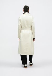 Dlhý béžový kabát so zaväzovaním v páse, zadným rozparkom a jedným radom gombíkov. Nosí sa cez čierne nohavice a topánky s kontrastnými bielymi prvkami.