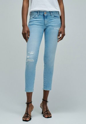 Mujer con vaqueros pitillo cortos y desgarrados de color azul claro y sandalias negras de tiras con tacón alto, posando frente a un fondo liso.