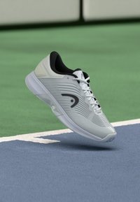 Sapatilha desportiva branca e preta pairando acima de um court de ténis azul e verde com uma linha de contorno branca.