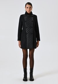 Cappotto nero con motivo a rombi imbottito sul davanti, bottoni doppi, colletto alto e due tasche laterali. Indossato su collant neri e stivali.