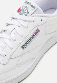 Bijela Reebok Classic tenisica s glatkom kožnom gornjom stranom, ravnim vezicama, srebrnim detaljima i logotipskim oznakama s dizajnom britanske zastave.