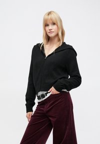 Μαύρο κοντό φούτερ με φερμουάρ και κουκούλα, πλεκτή υφή και τσέπες μπροστά, συνδυασμένο με μπορντό παντελόνι κορντόνι και λευκό πουκάμισο με βολάν από κάτω.