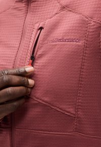 Κοντινή λήψη ενός χεριού που τραβάει το φερμουάρ σε ένα υφασμάτινο μοβ μπουφάν με μικρή τσέπη στο στήθος με φερμουάρ και κεντημένο λογότυπο.