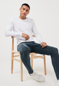 Hvid langærmet skjorte kombineret med blå denimjeans og hvide sneakers, siddende på en træstol med et vævet sæde.