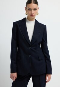 Námořnický blazer s proužkem vyrobený z upravené tkaniny, má dva přední knoflíky, klopové límce a dvě boční kapsy. Hladký povrch.