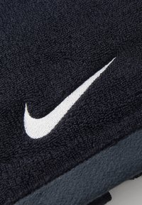 Sort håndklæde i tekstureret mikrofiberstof med et fremtrædende hvidt Nike Swoosh-logo syet på. Kantene er pænt færdiggjorte.