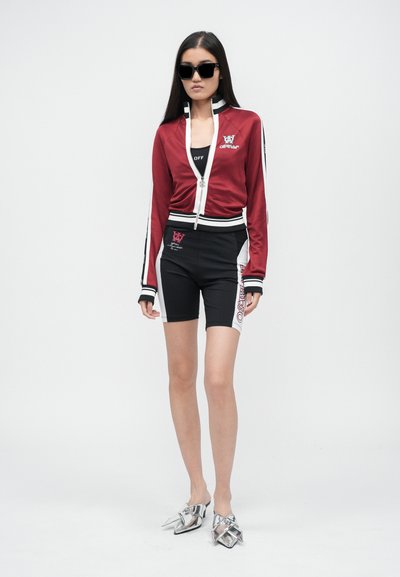 Giacca rossa con strisce bianche e logo, pantaloncini neri con grafiche laterali e scarpe metalliche con punta a punta; il modello indossa occhiali da sole neri.