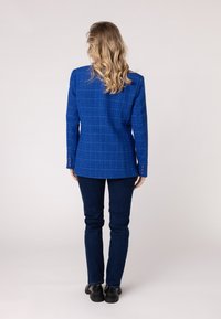 Modrý textúrovaný blazer s kockovaným vzorom, s dvoma gombíkmi na rukávoch, skombinovaný s tmavými džínsami a čiernymi topánkami. Pohľad zozadu.