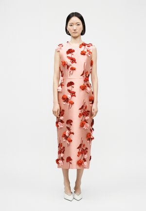 Vrouw staand, gekleed in een mouwloze roze midi-jurk bedekt met rode en roze 3D bloemapplicaties en witte pumps met een puntige neus.