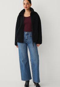 Zwarte fleece zip-up jack met hoge kraag, gecombineerd met een bordeauxkleurige top en lichtblauwe wijde jeans. Geplaatst tegen een neutrale achtergrond.