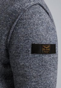 Gray-blue knit sweater fabric with a textured surface, featuring a black woven label on the sleeve with gold text.