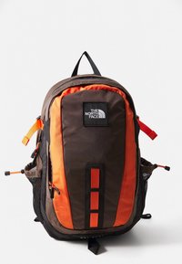Hnědý a oranžový batoh s přední kapsou na zip, síťovanými bočními kapsami a nastavitelnými popruhy. Má logo a reflexní prvky.