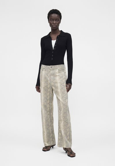 Γυναίκα που στέκεται, φοράει ένα μαύρο μπλουζάκι με μακριά μανίκια και κουμπιά, μπεζ παντελόνια με φαρδιά πόδια και σχέδιο φίδι, και μαύρα ανοιχτά πέδιλα, σε λευκό φόντο.