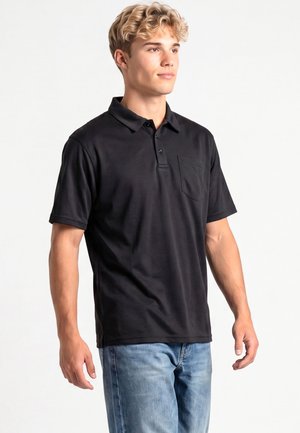 Jeune homme aux cheveux blonds bouclés portant un polo noir à manches courtes et un jean bleu, debout et regardant légèrement vers la droite.