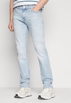 Homme portant un jean slim bleu clair et des baskets blanches, debout avec une main dans la poche devant un fond uni.