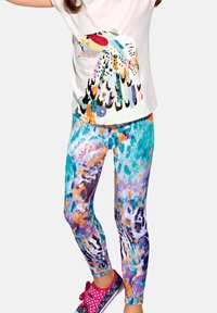 Dziecko ubrane w biały top z kolorową grafiką ptaka, wielobarwne abstrakcyjne legginsy oraz różowe trampki w groszki, stojące z podniesionymi ramionami.