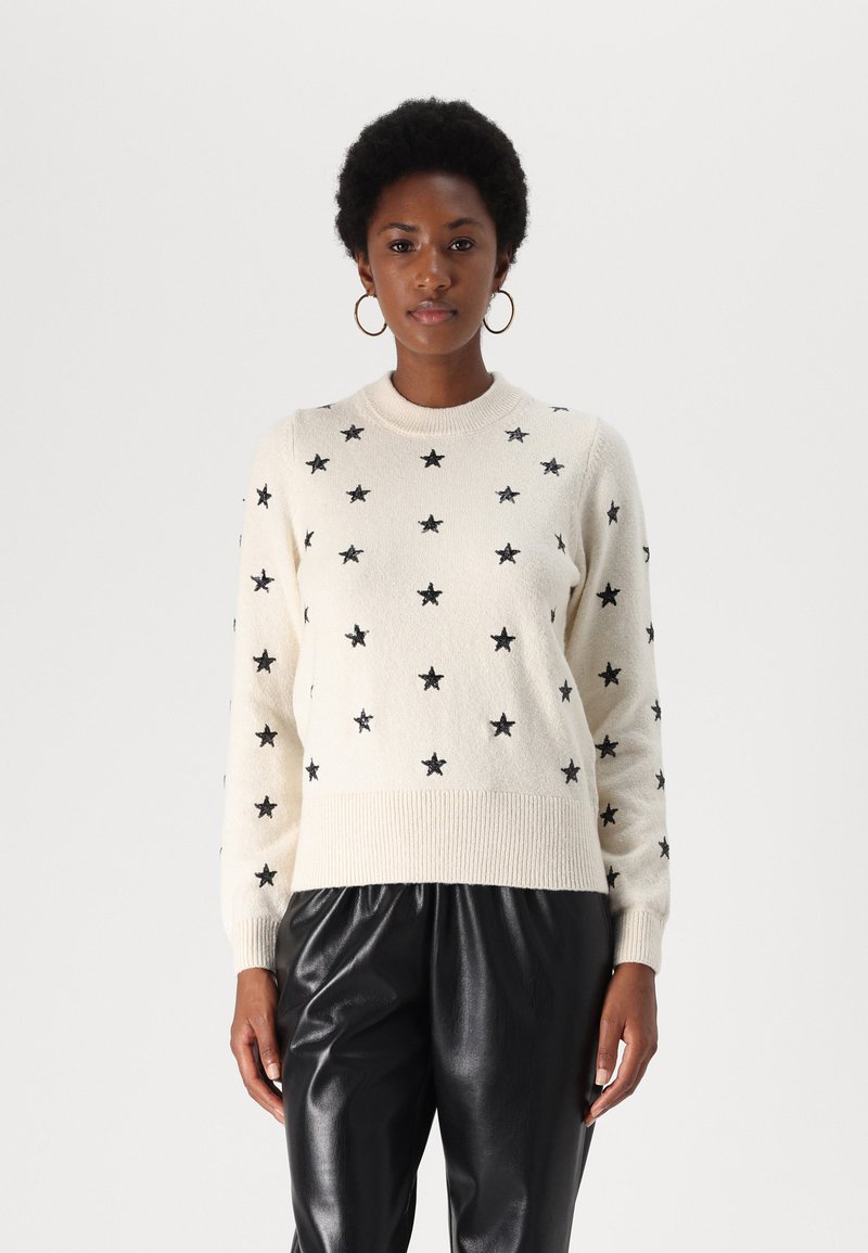 Pull en tricot crème avec des motifs d'étoiles noires, col et poignets côtelés. Associé à un pantalon en cuir noir.