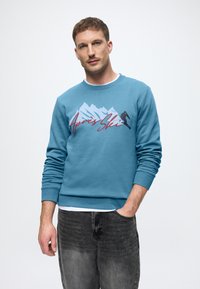 Hanorac turcoaz cu grafică montană și text "Après Ski", guler rotund, manșete ribbate și textură moale. Purtați cu blugi închiși la culoare.