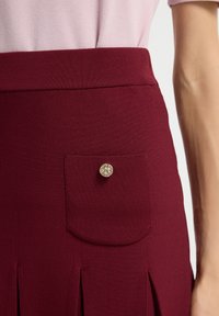 Bordeaux rok met een geribbeld patroon, voorzien van een klein voorvak en een metalen knoopaccent. Uitlopende vorm met plooien aan de zoom.