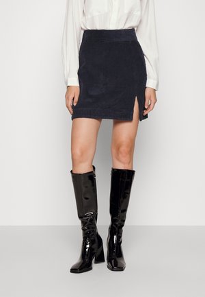 Vrouw draagt een marineblauwe corduroy minirok met een voorste split, een wit blouse met lange mouwen en zwarte, leren, kniehoge laarzen met hak.