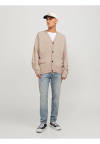 Cardigan lavorato a maglia beige con scollo a V, tre grandi bottoni, polsini a coste, abbinato a jeans azzurri chiari e sneaker nere. Cappellino con logo.
