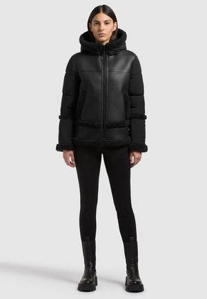 Femme portant une doudoune noire à capuche avec bordure en polaire, pantalon noir ajusté et bottes noires, debout devant un fond uni gris clair.