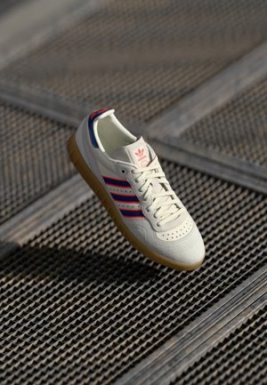 Valkoinen Adidas-tennari, jossa on punaiset ja laivastonsiniset raidat sekä kumipohja, sijoitettuna teksturoidulle metallipinnalle, jossa on diagonaalinen ruudukkomalli.