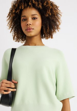 Νεαρή γυναίκα με φυσικά σγουρά μαλλιά που φοράει ανοιχτοπράσινο πουλόβερ, κρατώντας το λουράκι μιας μαύρης τσάντας ώμου μπροστά σε λευκό ουδέτερο φόντο.