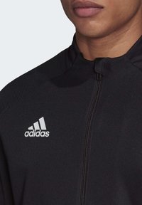 Zwarte ritsjack gemaakt van gladde stof, met een hoge kraag en een wit Adidas-logo op de linkerborst.