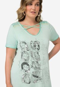 Camiseta verde menta de manga corta, escote en V, con acentos entrelazados en los hombros y diseño de texto gráfico con lentejuelas plateadas y texturas ligeras.