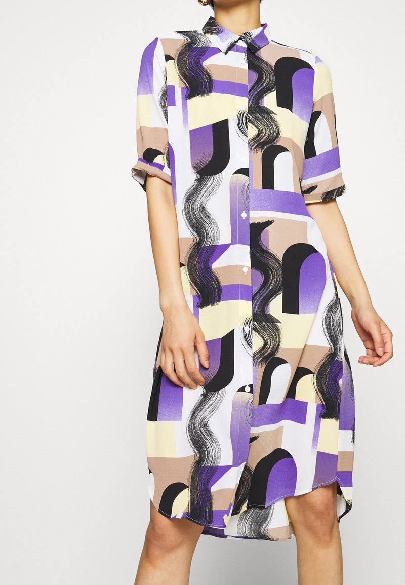 Femme portant une robe à manches courtes et à boutons avec un motif géométrique abstrait dans des tons de violet, beige, noir et crème.