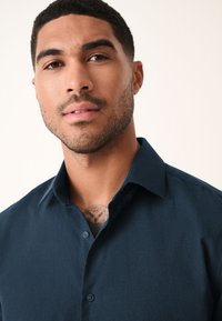 Donkergroen-blauw overhemd met knopen, voorzien van een gestructureerde kraag en een gladde textuur, gedragen door een model met een zichtbare tatoeage op de borst.