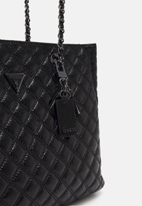 Czarna pikowana torba na ramię o błyszczącym wykończeniu, z łańcuszkowymi paskami i metką z logo. Posiada trójkątną plakietkę z logo oraz fakturowany skórzany element.