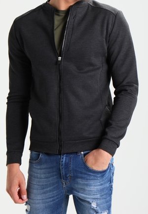 Homme portant une veste zippée gris foncé par-dessus une chemise vert olive avec un jean bleu déchiré, debout avec une main dans la poche.