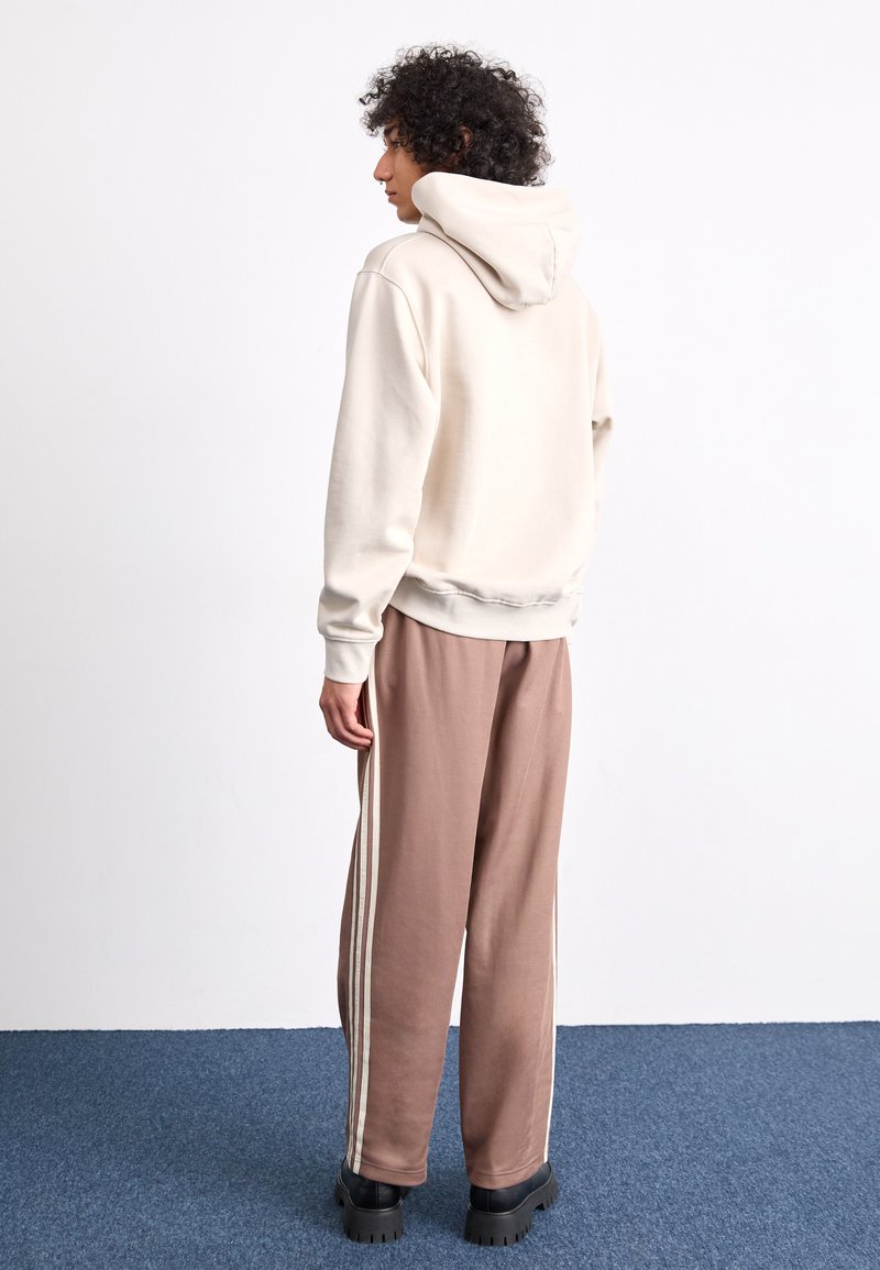 Felpa crema con un ampio cappuccio, abbinata a pantaloni sportivi marroni con strisce bianche laterali. Scarpe platform nere spesse completano l'outfit.