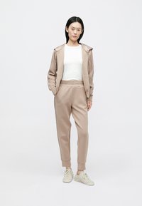 Ανοιχτό μπεζ φόρμα από μαλακό ύφασμα, με μπουφάν με φερμουάρ και στενόμακρα παντελόνια με μανσέτες. Το μοντέλο φοράει αθλητικά παπούτσια σε ανοιχτό χρώμα.